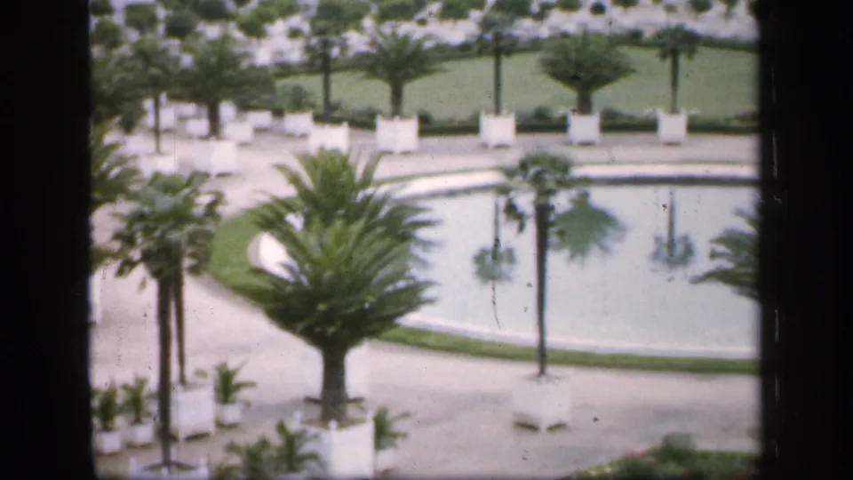 1949 France home-movie still showing the lower part of a formal garden with a large palm, pool edge, paths, white planters, and trimmed green lawns.