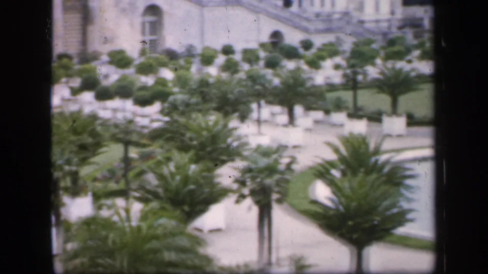 1949 France frame showing a formal landscaped garden with palms, white planters, a curved pool, paths, and part of a substantial stone building.