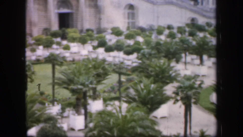 1949 France home-movie frame showing a formal garden with small palmetto trees, clipped shrubs, paths, and a pale classical facade in soft focus.