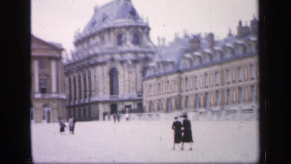 1949 France home-movie view of a broad courtyard bordered by ornate stone buildings, with a few distant visitors walking across the open space.