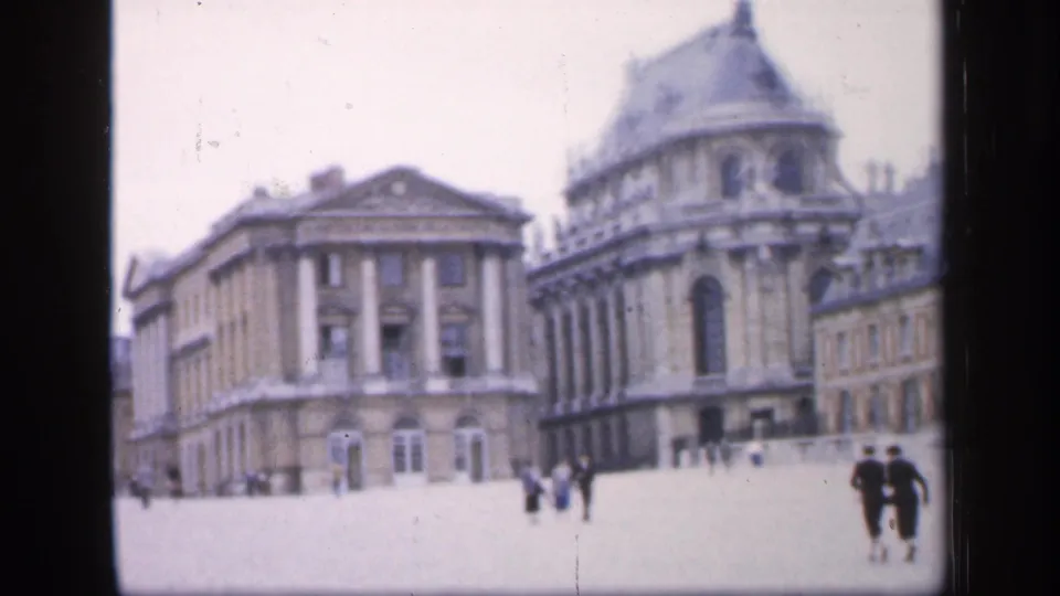 1949 France frame showing a broad courtyard, a columned classical building, an ornate mansard-roofed wing, and distant visitors walking nearby.