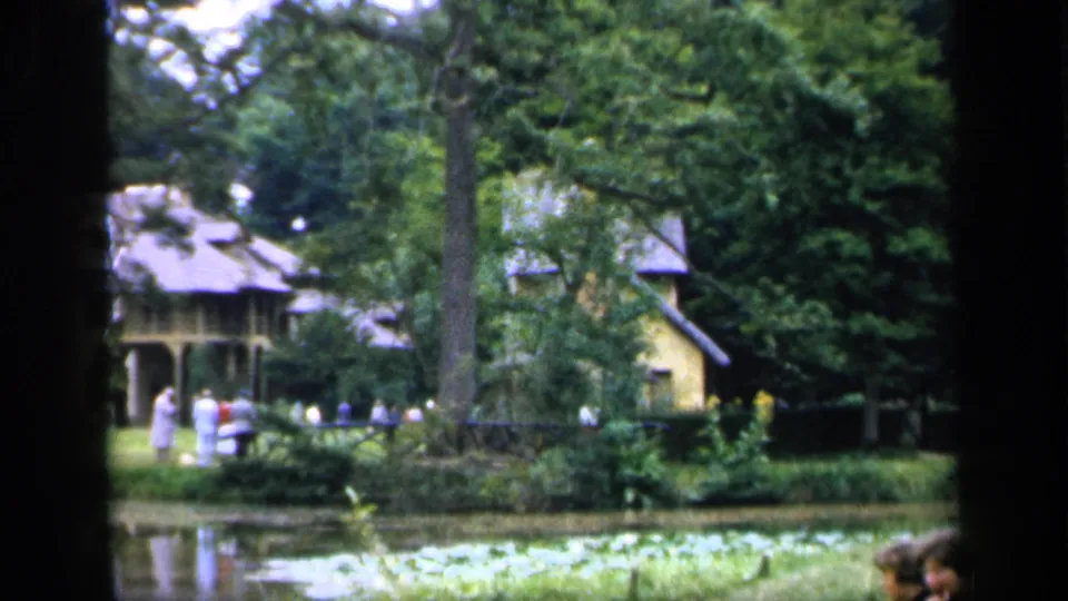 1949 France home-movie garden scene with a pond, lily pads, trees, a yellow house, and distant visitors walking near the water.