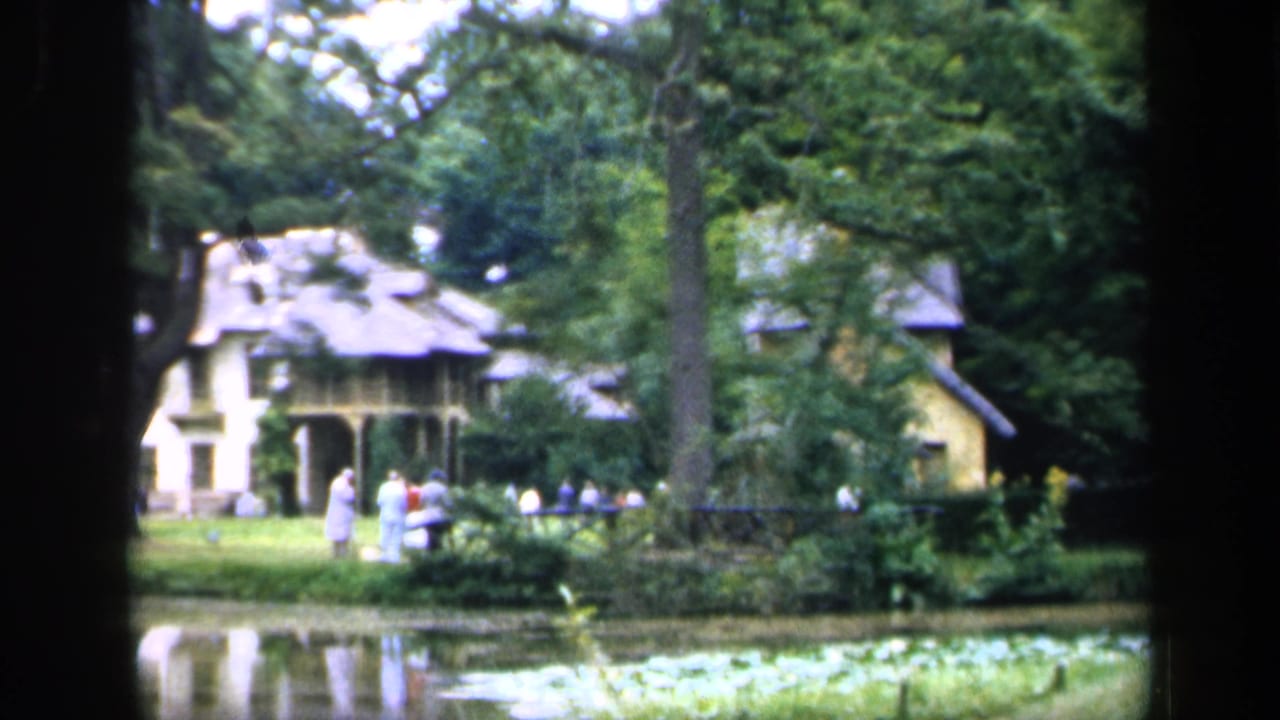 Large house beyond a pond, with trees, lawn, and distant people walking.