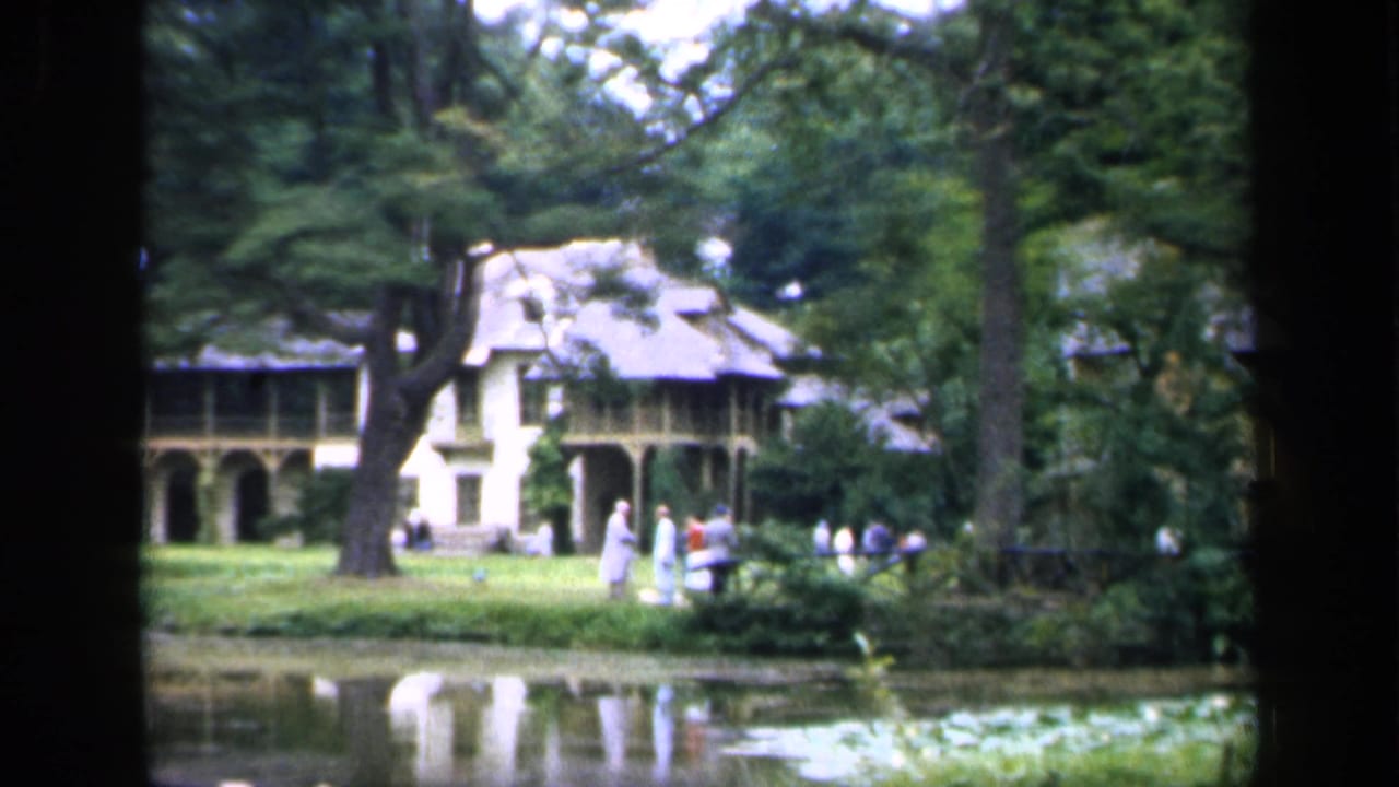 House and porch seen through trees, with people standing near the lawn and pond.