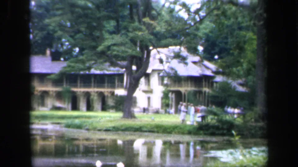 1949 France 8mm frame showing people strolling near a pond, with a large house, arched porch, lawn, and heavy tree cover behind them.