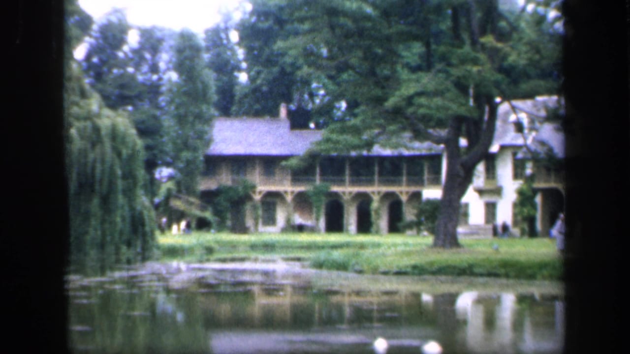 1949 8mm view in France of a long estate building with arched colonnade, lawn, trees, pond, and faint figures near the structure.
