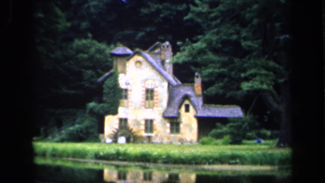 1949 8mm view in France of an ivy-covered stone cottage or small mansion across water, backed by dense trees and a grassy bank.