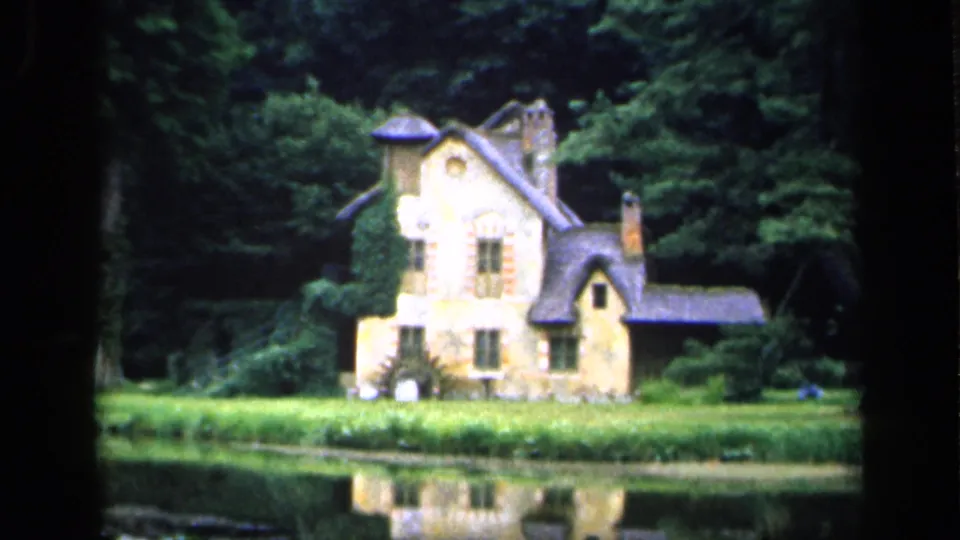 A French 1949 home-movie frame shows the ivy-covered stone cottage, low grass bank, surrounding trees, and a soft reflection in the pond.