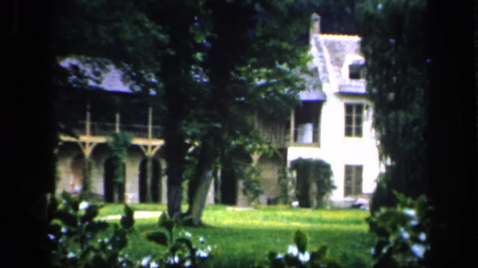 1949 France 8mm frame of a chateau facade with arched colonnades, a pale side wing, lawn, and trees partly obscuring the view.