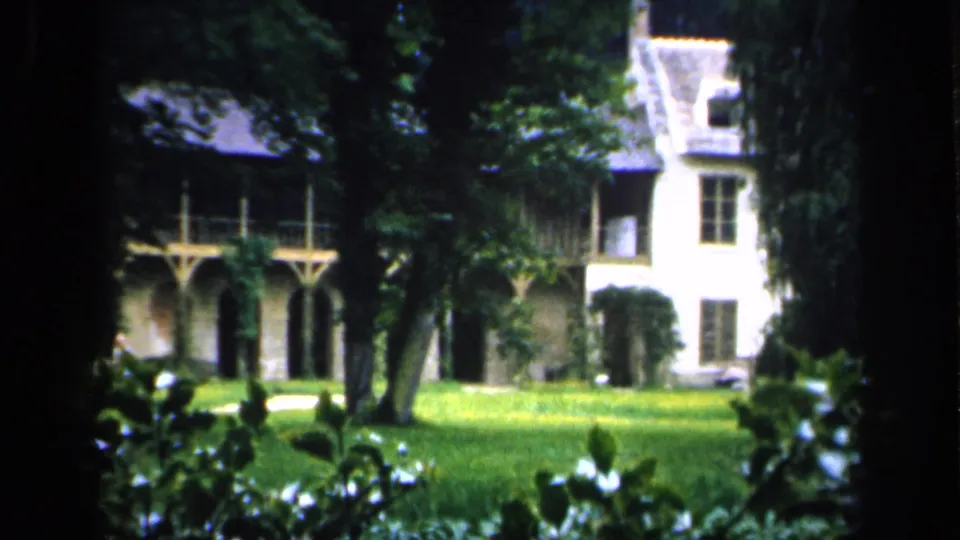 1949 France 8mm frame showing the chateau's arched porch and pale side section, with lawn and leafy foreground plants.