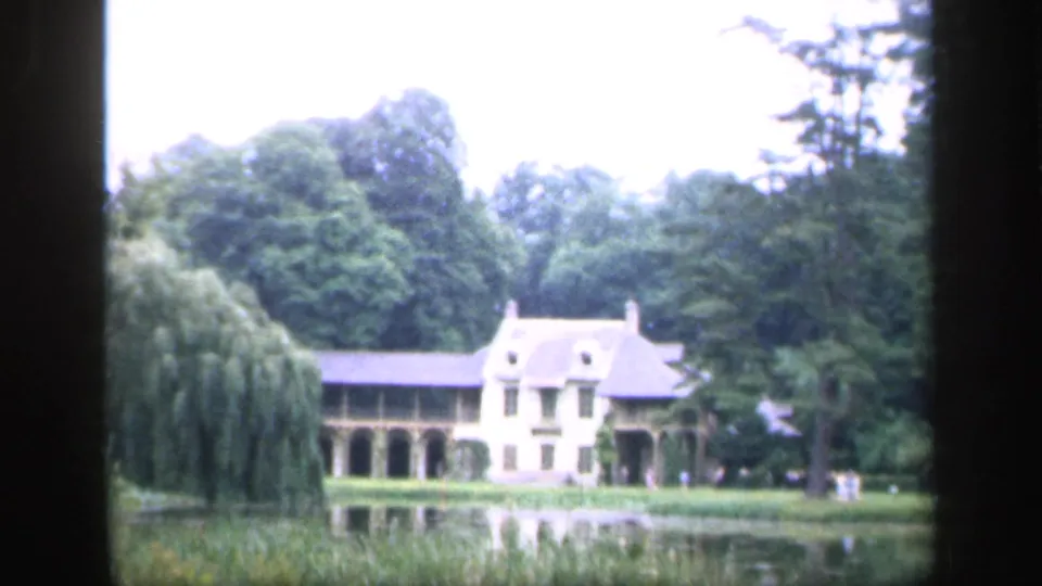 1949 France 8mm frame showing a pale chateau across a pond, with arched colonnade, slate roof, willow, and dense trees.