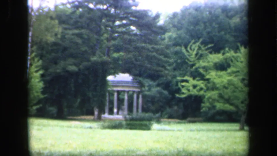 1949 France 8mm frame showing a small classical garden pavilion set among trees and shrubs beside an open lawn.