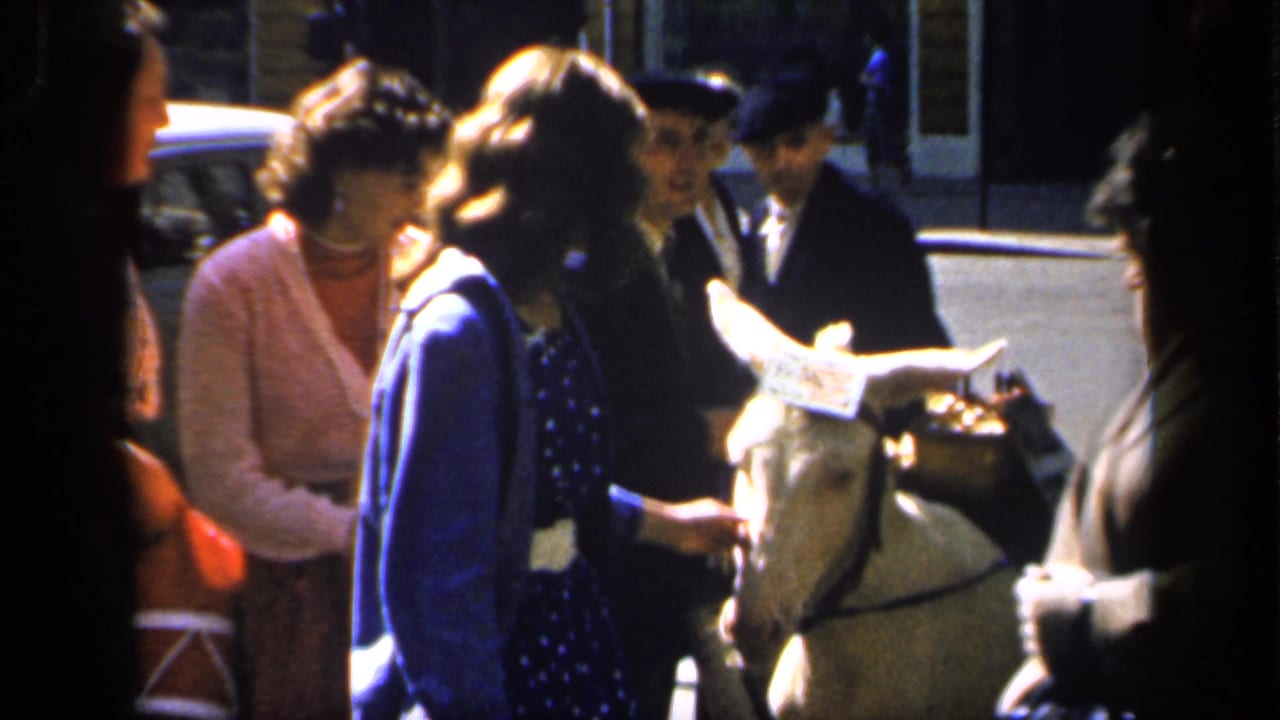 1949 France home-movie frame showing women and uniformed men near a small white donkey on a town street.