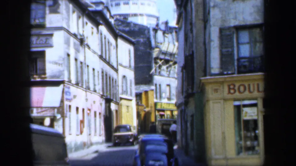 1949 France home-movie view down a narrow street with older buildings, parked cars, signage and a distant domed form.