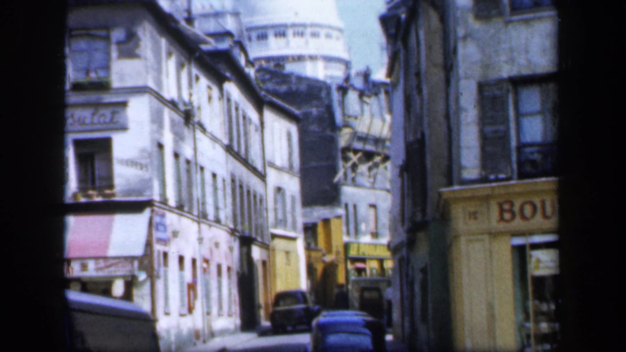 1949 8mm view down a Montmartre street with aging facades, parked cars, storefront signs, and Sacré-Cœur rising beyond the rooftops.