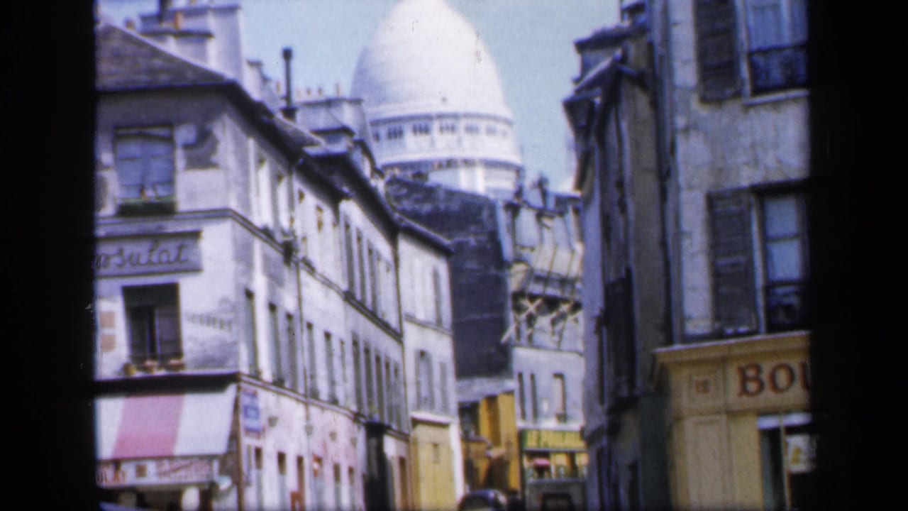 1949 Paris street frame showing shuttered windows, shop signage, parked cars, and the white Sacré-Cœur dome beyond Montmartre rooftops.