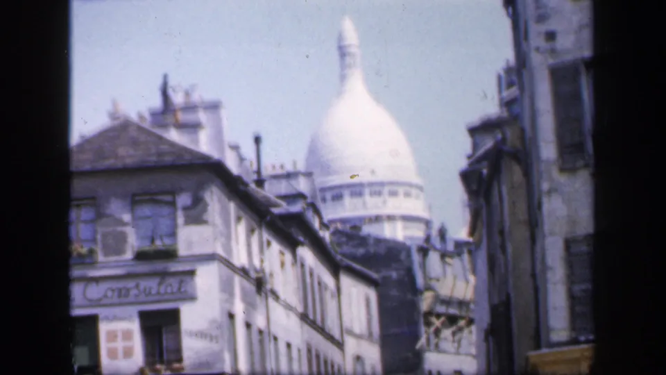1949 8mm frame of a Montmartre street with the Consulat sign on a corner facade and Sacré-Cœur visible over the roofline.