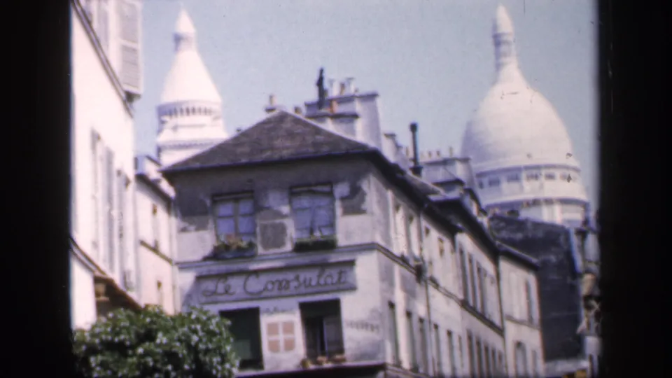1949 France home-movie frame showing Le Consulat signage and a Sacre-Coeur dome behind narrow Montmartre buildings.
