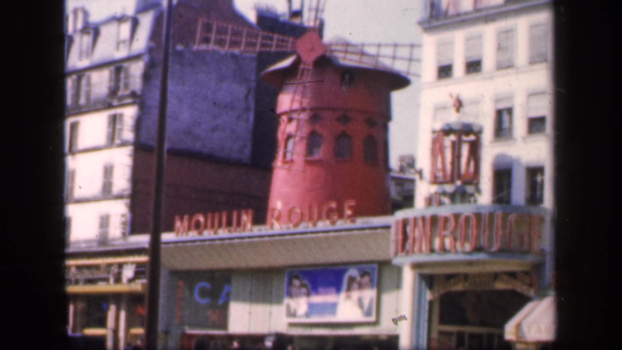 Moulin Rouge facade and entrance sign framed by buildings, with pedestrians partly visible along the sidewalk.