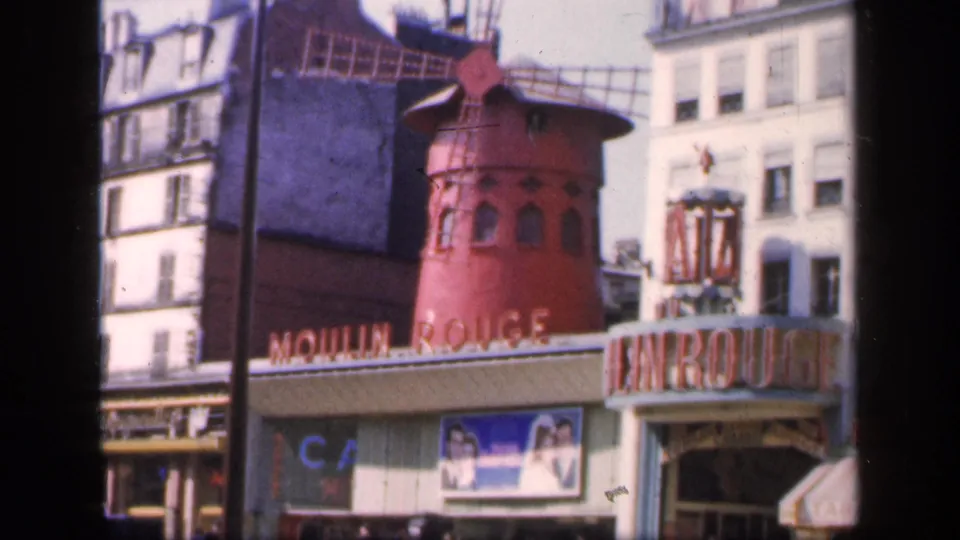Paris Moulin Rouge exterior on 8mm film, showing the red windmill, cabaret entrance sign, surrounding buildings, and passing street figures.