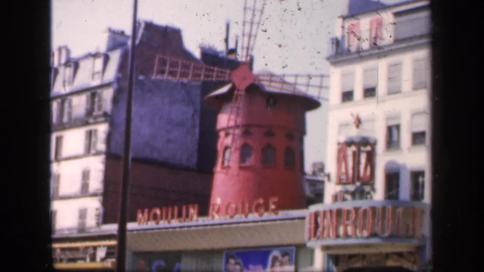 Vintage 8mm view of the Moulin Rouge in Paris, showing the red windmill, large entrance lettering, nearby facades, and people moving along the street.