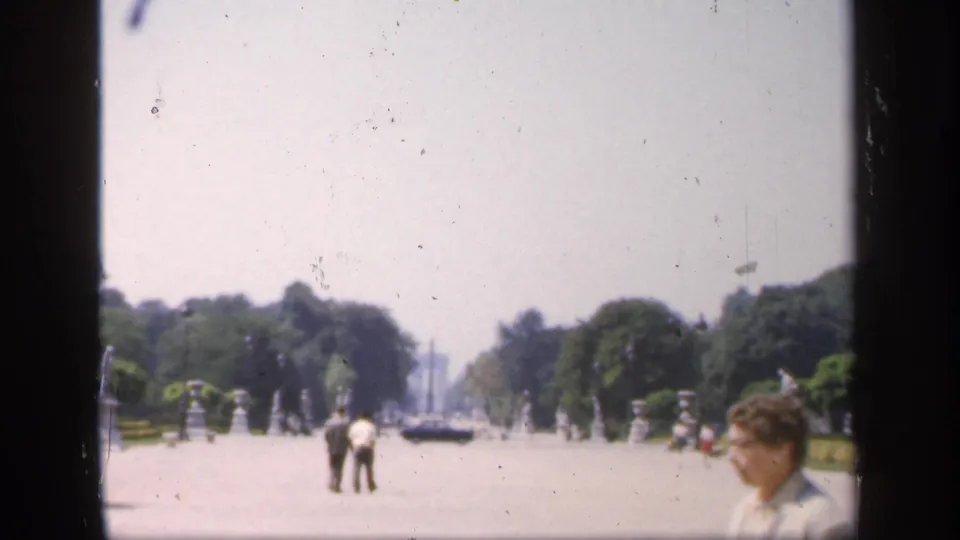 Wide 1949 France home-movie view of a sunlit public courtyard or avenue with pedestrians, trees, cars, and a partial face at the edge.