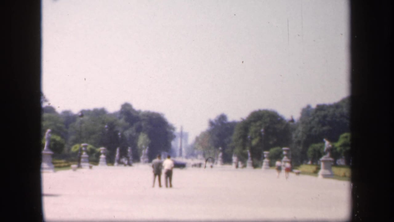 Open formal walkway with two people standing near the center and statues along tree-lined grounds.