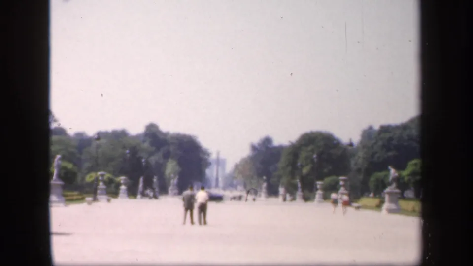 1949 France home-movie view down a wide formal walkway, with two visitors centered between trees, statues, and bright open grounds.