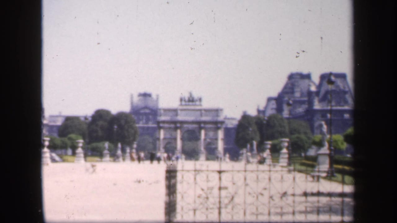 Wide courtyard view with arch, trees, rooftops, stonework, and small figures walking in the distance.
