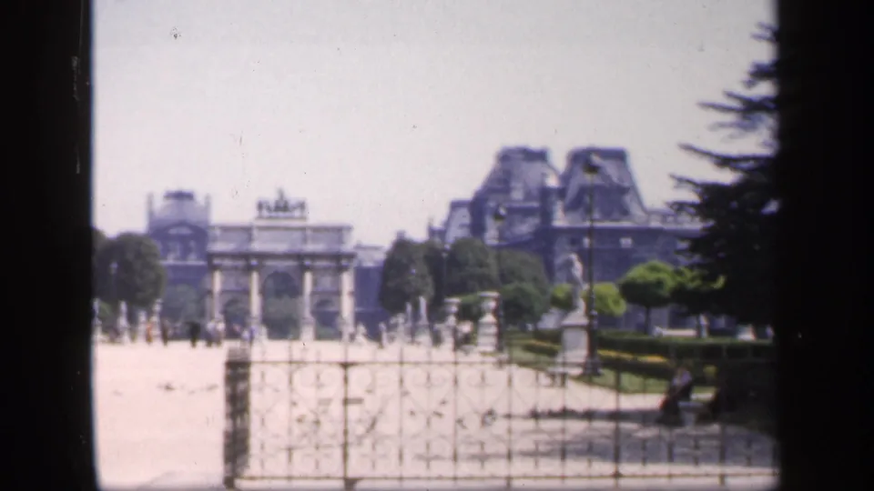 Soft 8mm view of a Paris landmark garden area in France, with an arch, stonework, rooftops, and pedestrians visible at a distance.
