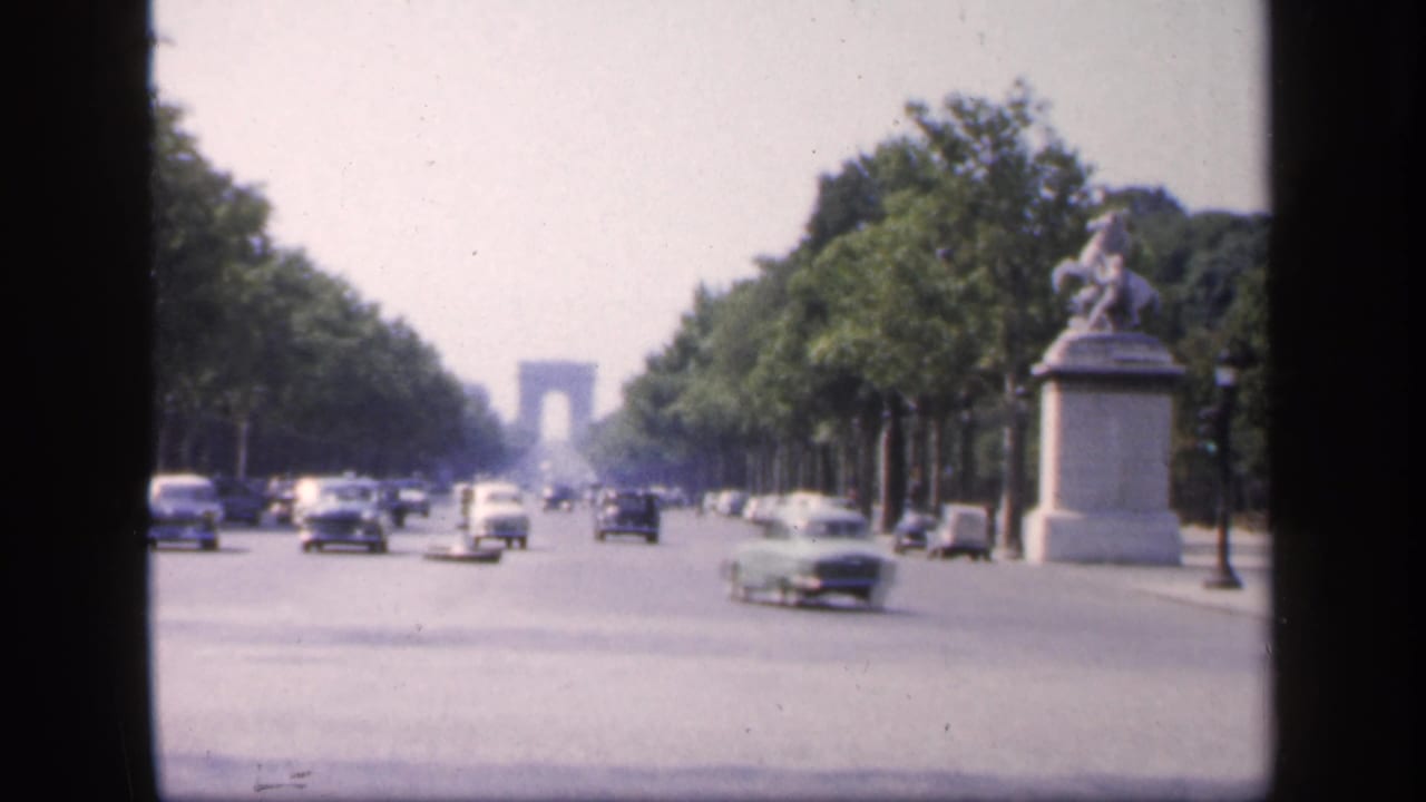 Tree-lined Paris boulevard with classic cars, distant Arc de Triomphe, and an equestrian statue at right.