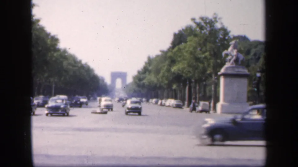 Paris boulevard traffic in 1949, showing several mid-century cars, rows of trees, a distant Arc de Triomphe, and a monument at the right edge.