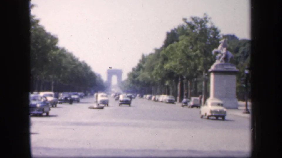1949 home-movie frame of a Paris boulevard, with classic automobiles crossing the foreground and the Arc de Triomphe softened by distance.