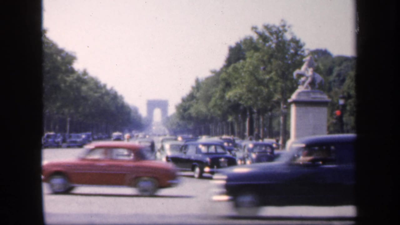 Midday 1949 Paris street traffic passes beneath trees, with a white statue pedestal nearby and the Arc de Triomphe beyond.
