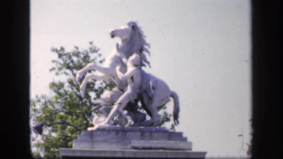 Soft 8mm view from 1949 France showing the white rearing horse sculpture on its stone base with trees in the background.