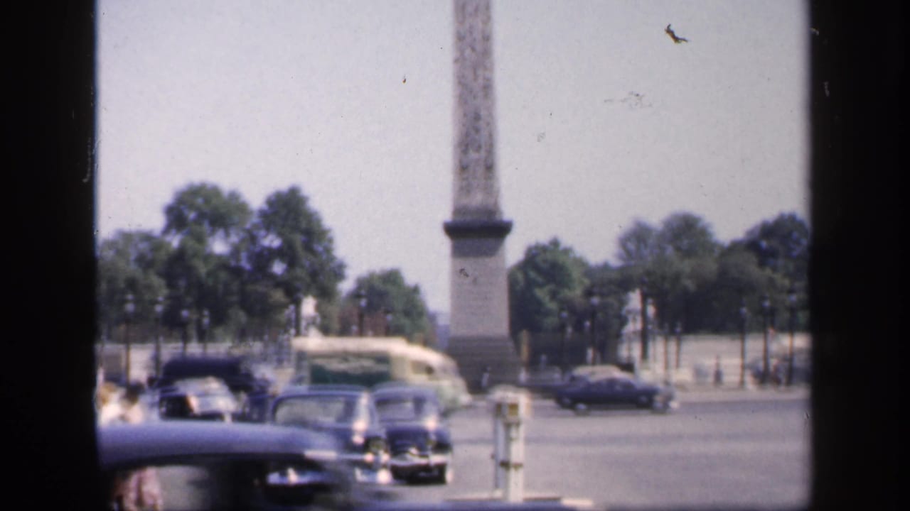 Classic cars near a tall stone monument in a tree-lined city square.