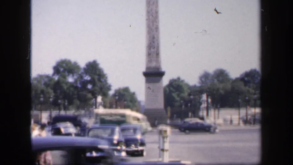 1949 France home-movie frame showing classic cars moving through a broad square beside a tall stone monument and trees.
