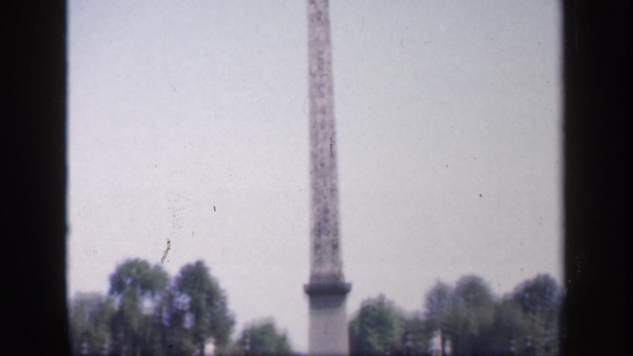 Soft 8mm view of a French city square, with a tall stone monument, trees, and mid-century cars in the distance.