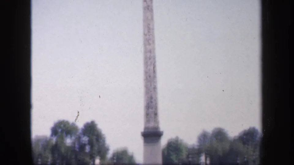 Soft 8mm view of a French city square, with a tall stone monument, trees, and mid-century cars in the distance.