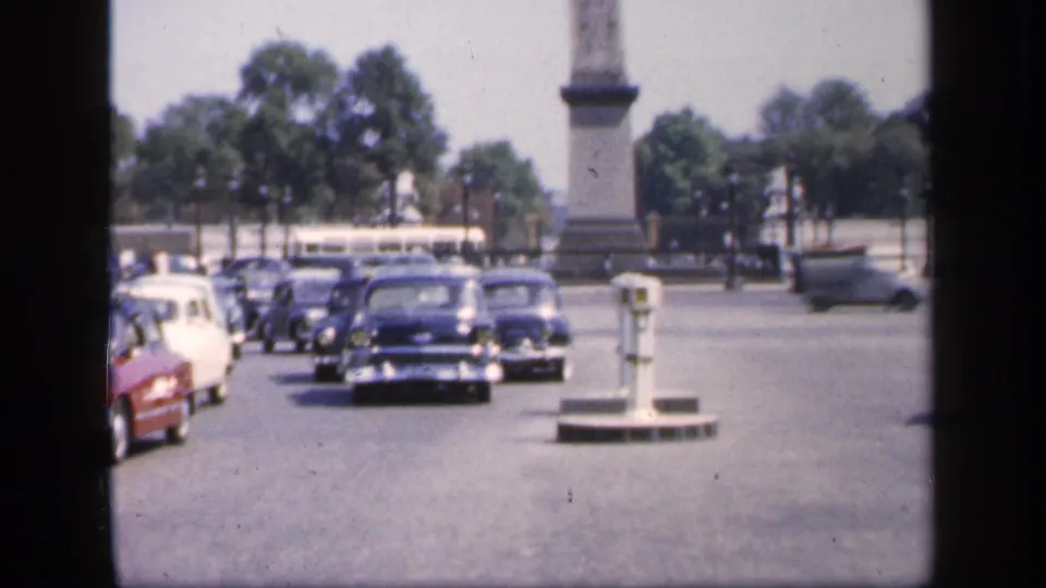 1949 France street view with classic cars, a bus, traffic posts, trees, and a stone monument at the edge of the frame.