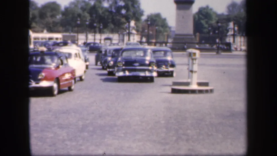 1949 France home-movie frame showing a red car, dark cars, a white car, traffic posts, trees, and a tall stone monument.