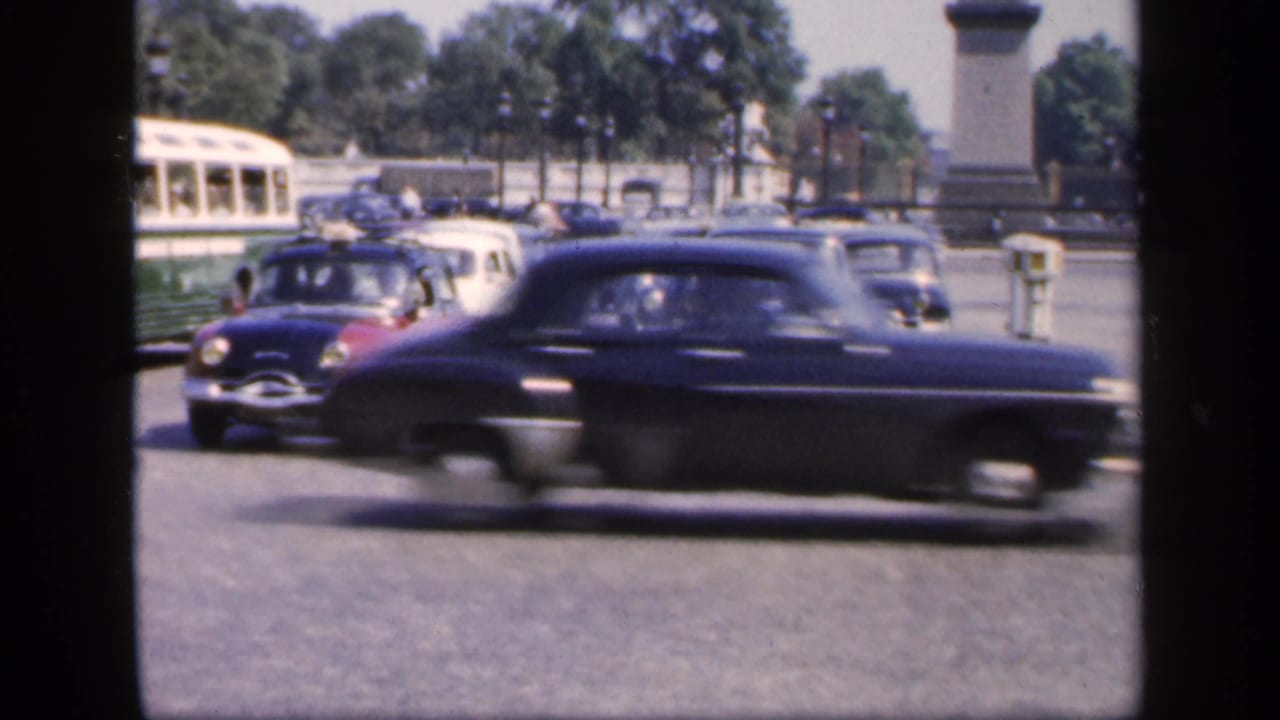 Vintage cars moving across a wide boulevard with trees and stone columns in the background.