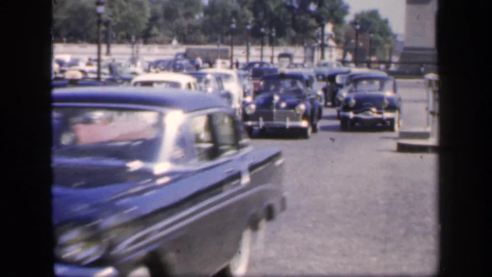 1949 France traffic frame with a blue mid-century car blurred in the foreground and several rounded cars approaching in the distance.