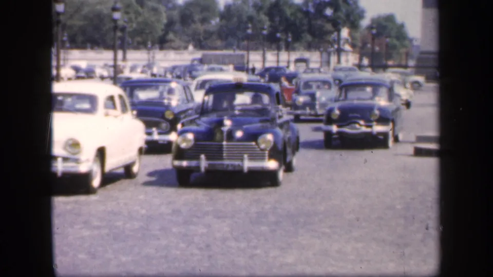 1949 France home-movie view of multiple mid-century cars driving toward the camera on a wide boulevard, with trees and distant people behind.