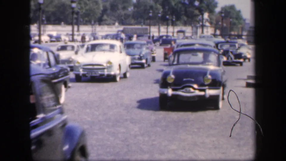 1949 France frame showing mixed mid-century cars moving along a wide boulevard, including a centered white car and a dark rounded car at right.
