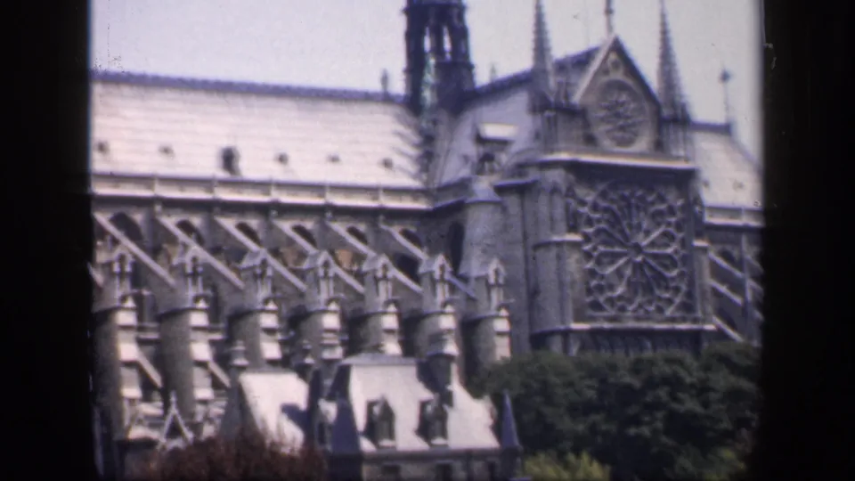 1949 France home-movie frame showing Notre-Dame Cathedral's rose window, Gothic buttresses, roofline, and trees from a slightly distant viewpoint.