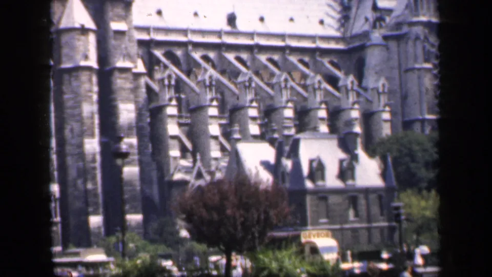 1949 France home-movie frame centered on Notre-Dame's Gothic buttresses and stonework, with trees, a foreground building, and street activity.