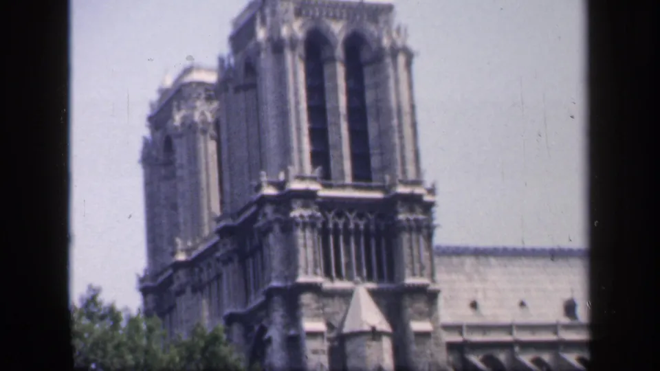 1949 France frame showing the upper portion of Notre-Dame Cathedral, with tower openings, stone ornament, roofline, and trees below.
