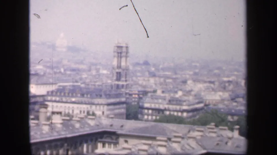 Soft 1949 8mm view over Paris rooftops, showing a tall tower structure and distant landmarks through haze.