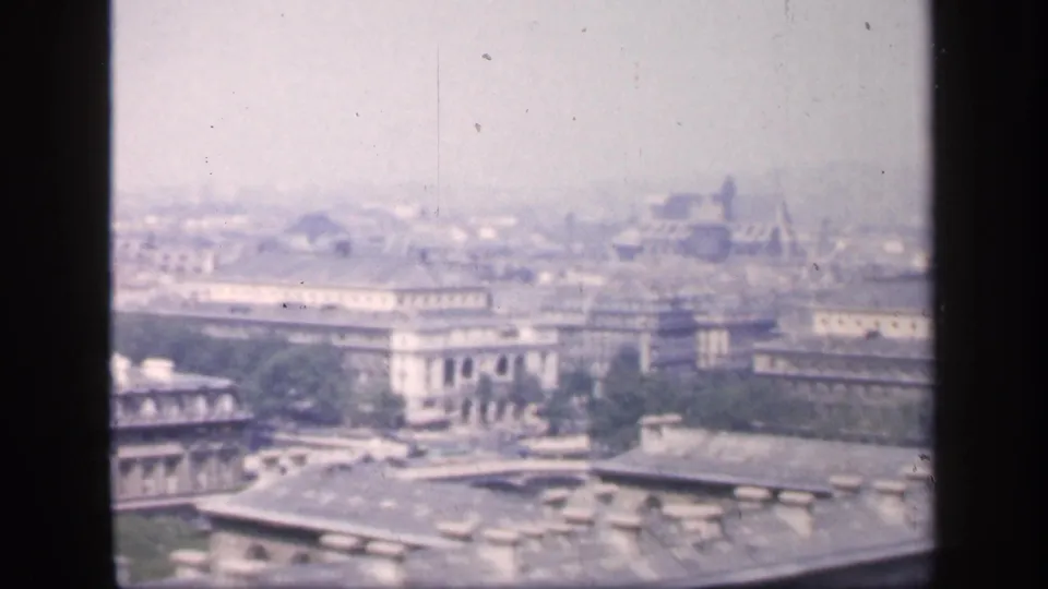 Hazy 1949 8mm frame of a French city seen from above, with rooftops, pale facades, trees, and distant structures.